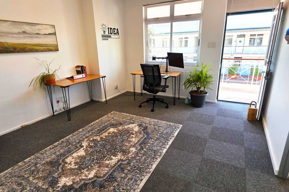 A bright office space featuring carpet tile flooring, large windows for natural light, and direct access to an outdoor balcony area.