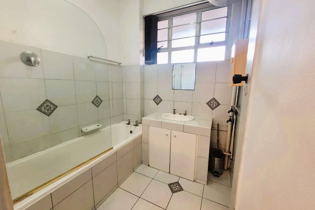 A dated bathroom featuring a built-in bathtub with a glass screen, a tiled vanity unit with a drop-in basin, and large windows.