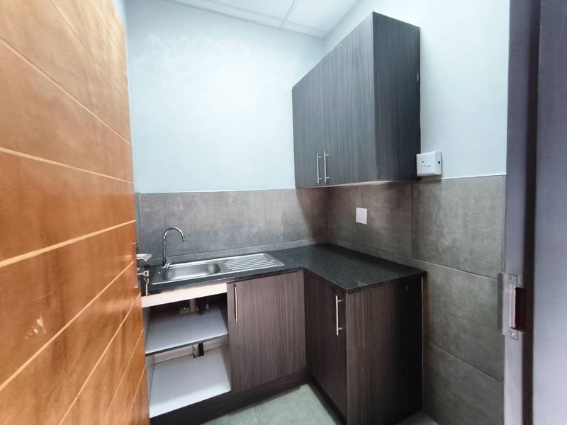 A compact, modern scullery area featuring dark wood-grain cabinetry, a stainless steel sink, granite countertops, and neutral wall tiling.