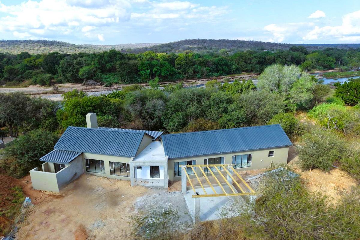 Aerial view of a residential property under construction, featuring a metal roof, large windows, and an unfinished patio area overlooking a river.