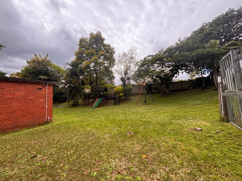A spacious backyard garden featuring a lawn area, mature trees, and perimeter boundary walls with a metal security gate.