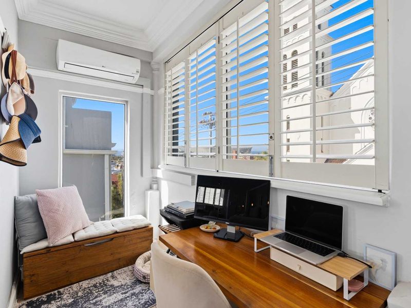 A bright home office featuring plantation shutters, decorative cornice, and a wall-mounted split-system air conditioner, offering a functional workspace with natural light.
