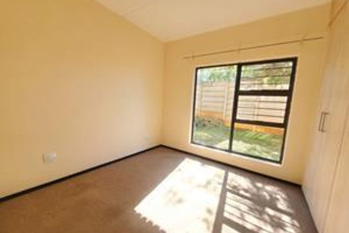 A bright, empty bedroom featuring neutral walls, carpeted flooring, and built-in wooden cupboards with a large window overlooking the garden.