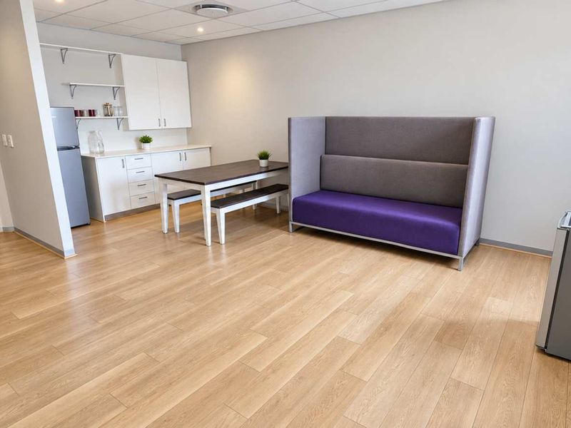 An open-plan office break area featuring light wood-look laminate flooring, a suspended ceiling with recessed lighting, and a compact kitchenette with built-in cabinetry.