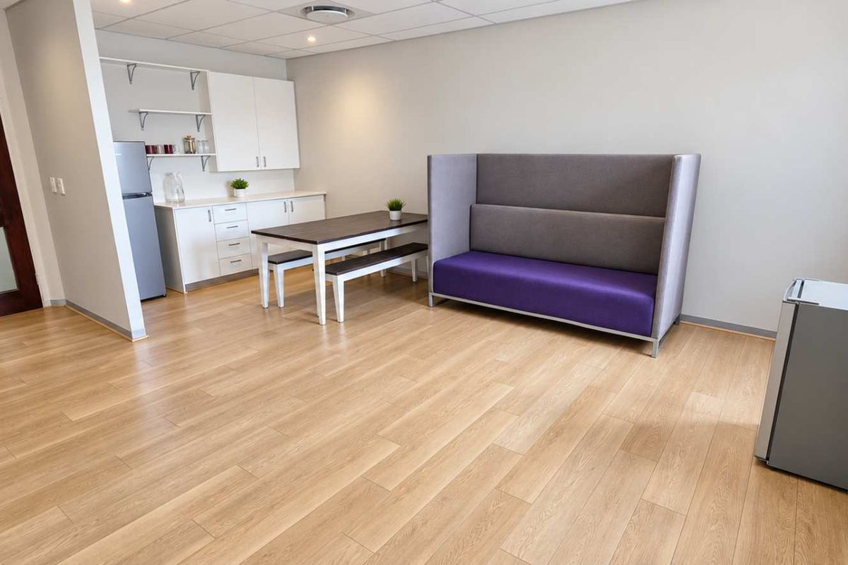 An open-plan office break area featuring light wood-look laminate flooring, a suspended ceiling with recessed lighting, and a compact kitchenette with built-in cabinetry.