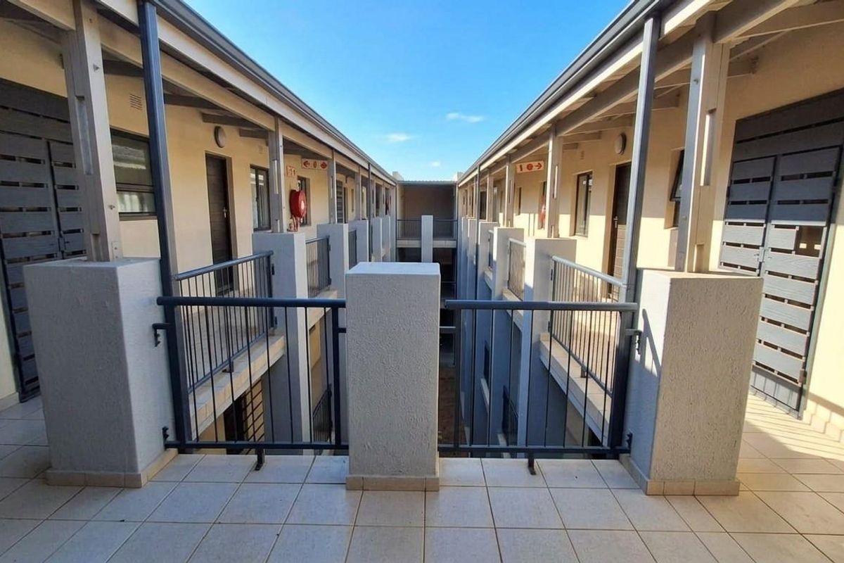 An exterior view of an apartment complex corridor featuring tiled walkways, metal balustrades, and access to multiple residential units.