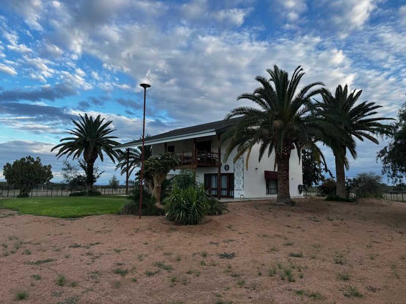 Rear view of a two-story farm residence featuring a timber balcony, sliding glass doors, and established palm trees set within a spacious rural landscape.