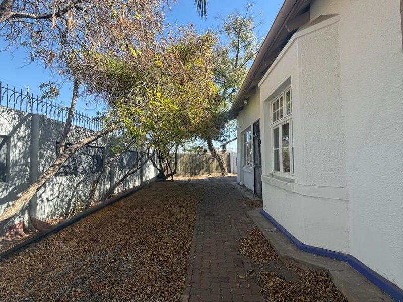 A side exterior view of the property featuring a paved walkway, perimeter wall with security spikes, and established trees along the boundary.
