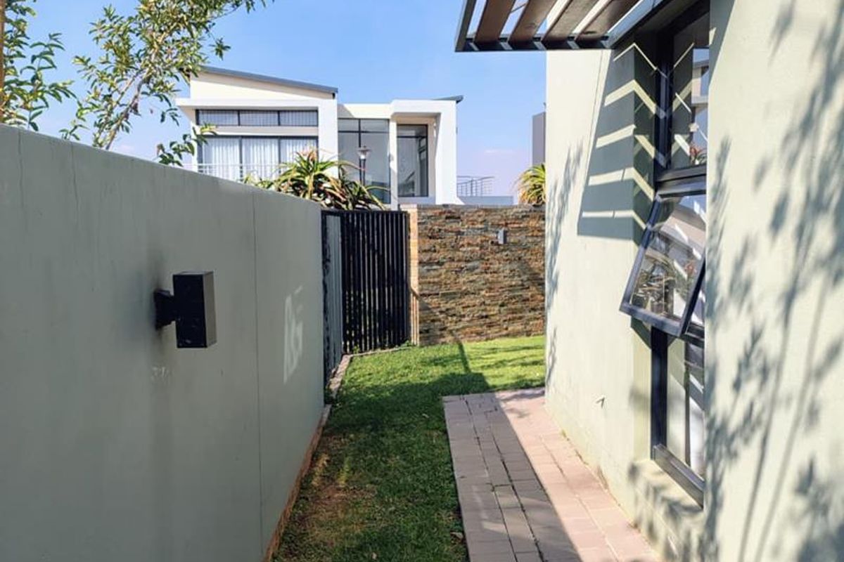 A side walkway featuring paved path, manicured lawn, boundary wall with exterior lighting, and a modern window with a fixed metal awning.
