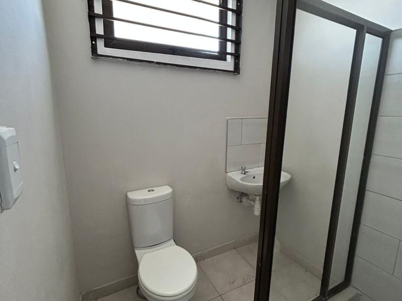 A functional bathroom featuring a toilet, wall-mounted basin, and a glass-enclosed shower with tiled finishes and security bars on the window.