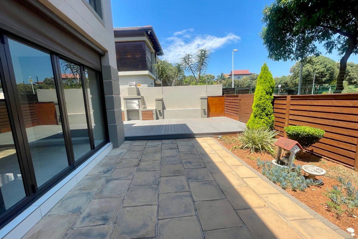 A well-maintained rear patio area featuring paved flooring, a raised composite deck, and a built-in outdoor braai, set within a landscaped garden.