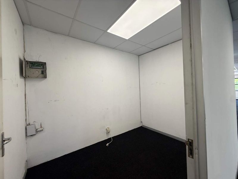 A small, empty office space featuring a suspended ceiling, recessed lighting, and dark carpeting, requiring cosmetic wall repairs and maintenance.