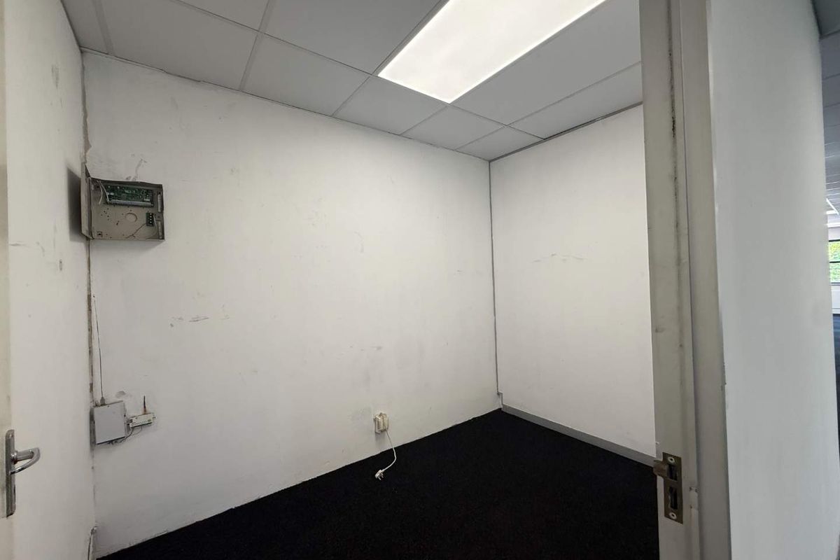 A small, empty office space featuring a suspended ceiling, recessed lighting, and dark carpeting, requiring cosmetic wall repairs and maintenance.