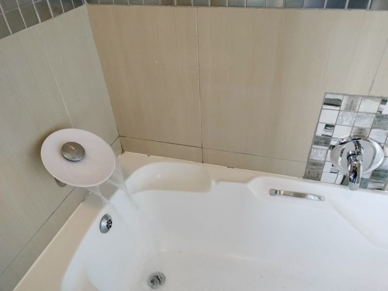 A close-up view of a built-in bathtub featuring a modern waterfall faucet, chrome mixer tap, and neutral ceramic wall tiles with mosaic accents.