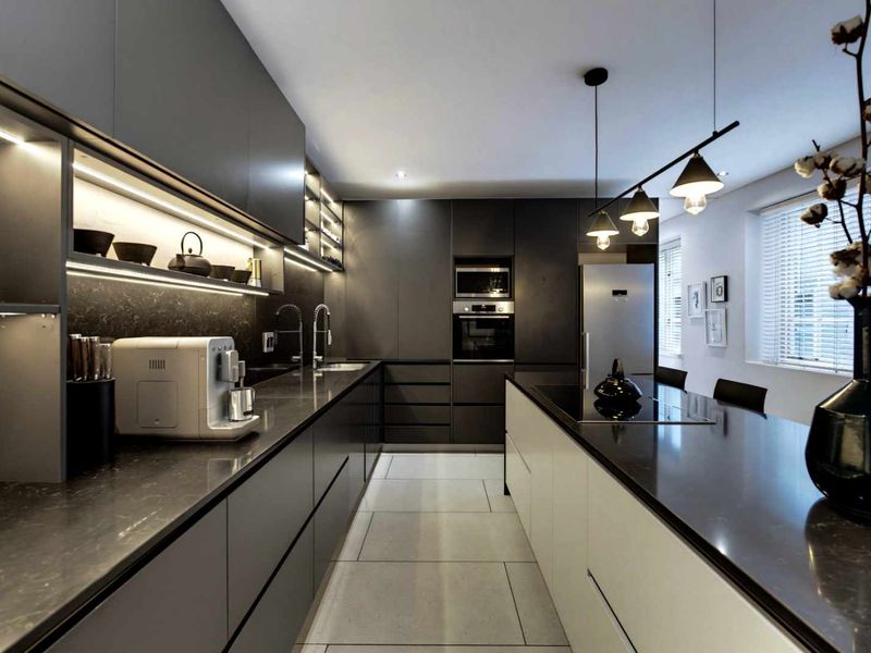 Modern, high-end kitchen featuring sleek dark cabinetry, stone countertops, integrated appliances, and a central island with a cooktop and pendant lighting.