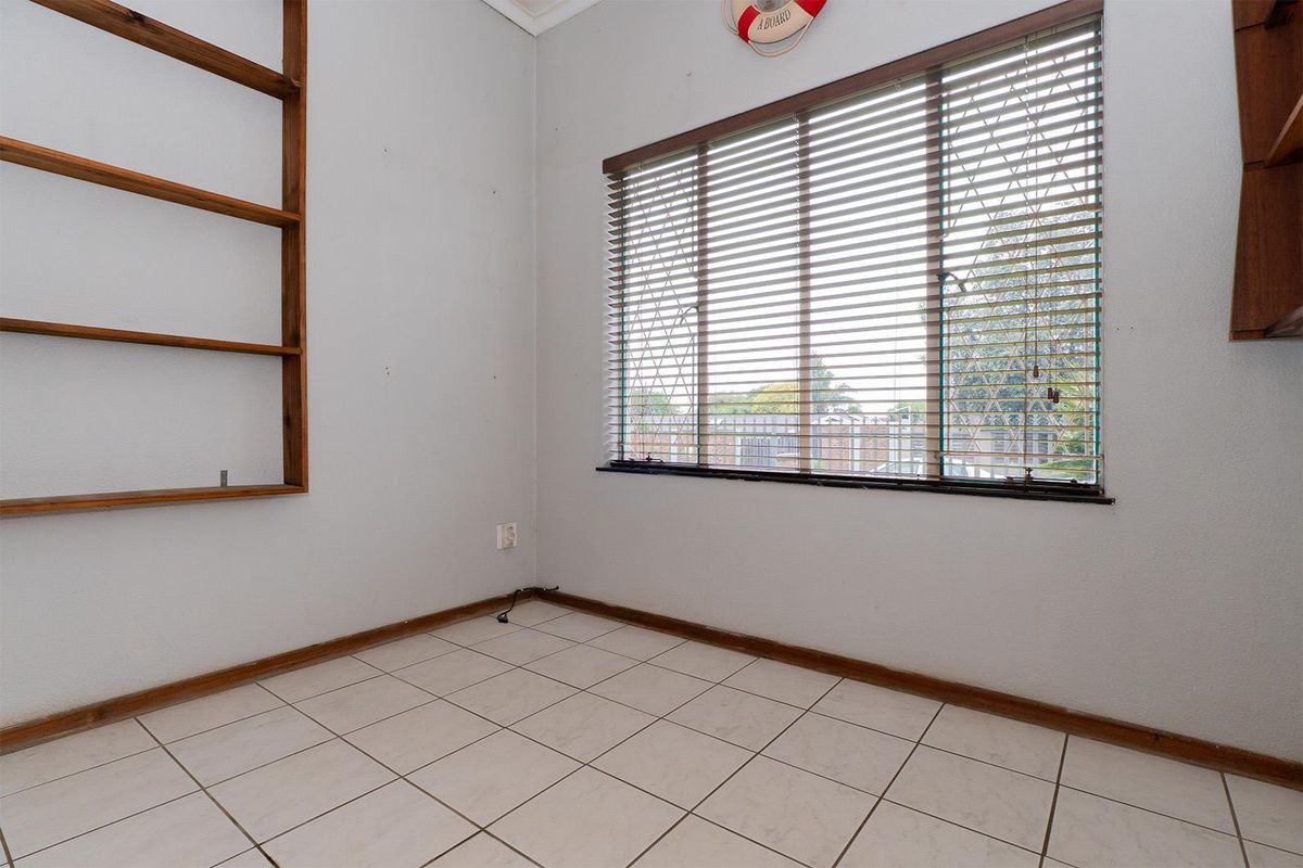 A functional bedroom or office space featuring tiled flooring, built-in wooden shelving, and a window with fitted blinds.