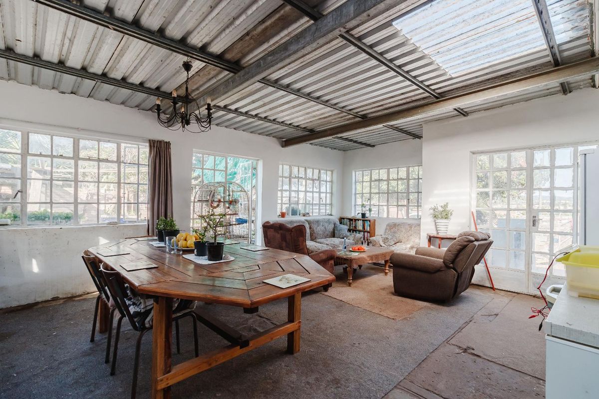 Spacious open-plan living and dining area featuring exposed steel beams, a corrugated metal roof with translucent panels, and large steel-framed windows.