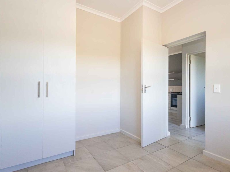 A clean, empty bedroom featuring neutral tiled flooring, a built-in wardrobe, and a view through to the adjacent kitchen area.