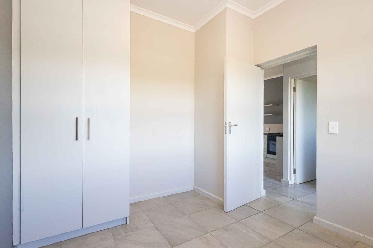 A clean, empty bedroom featuring neutral tiled flooring, a built-in wardrobe, and a view through to the adjacent kitchen area.