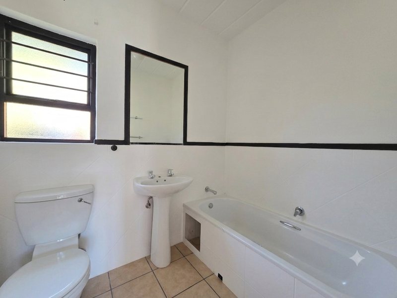 A functional bathroom featuring a pedestal basin, bathtub, and toilet, with tiled flooring and a window with security bars.