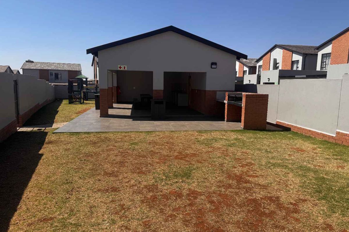 A spacious rear garden featuring a covered patio with a built-in braai, tiled flooring, and enclosed by brick boundary walls.