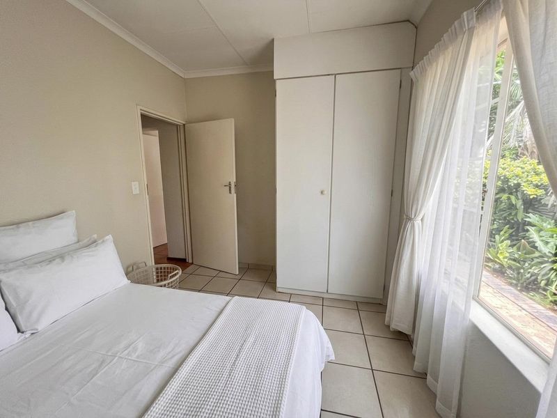 A functional bedroom featuring tiled flooring, a built-in wardrobe, and a large window providing natural light and garden views.