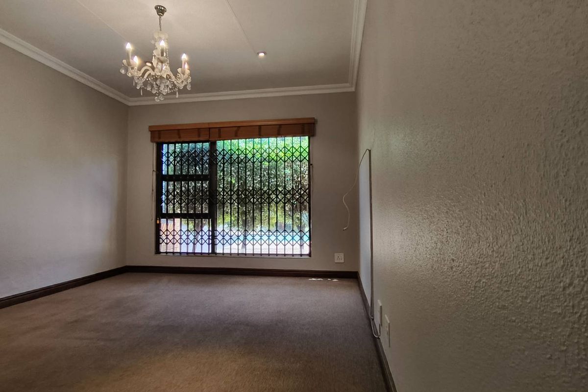 A spacious bedroom featuring neutral walls, carpeted flooring, a decorative chandelier, and a window fitted with security bars.