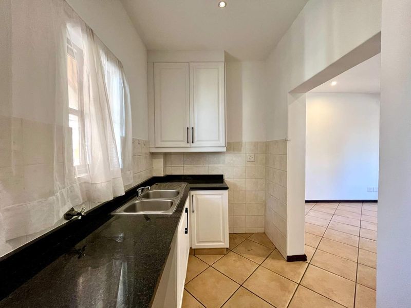 A functional kitchen featuring white cabinetry, dark granite countertops, a double basin sink, and neutral tiled flooring, leading into an adjacent living area.