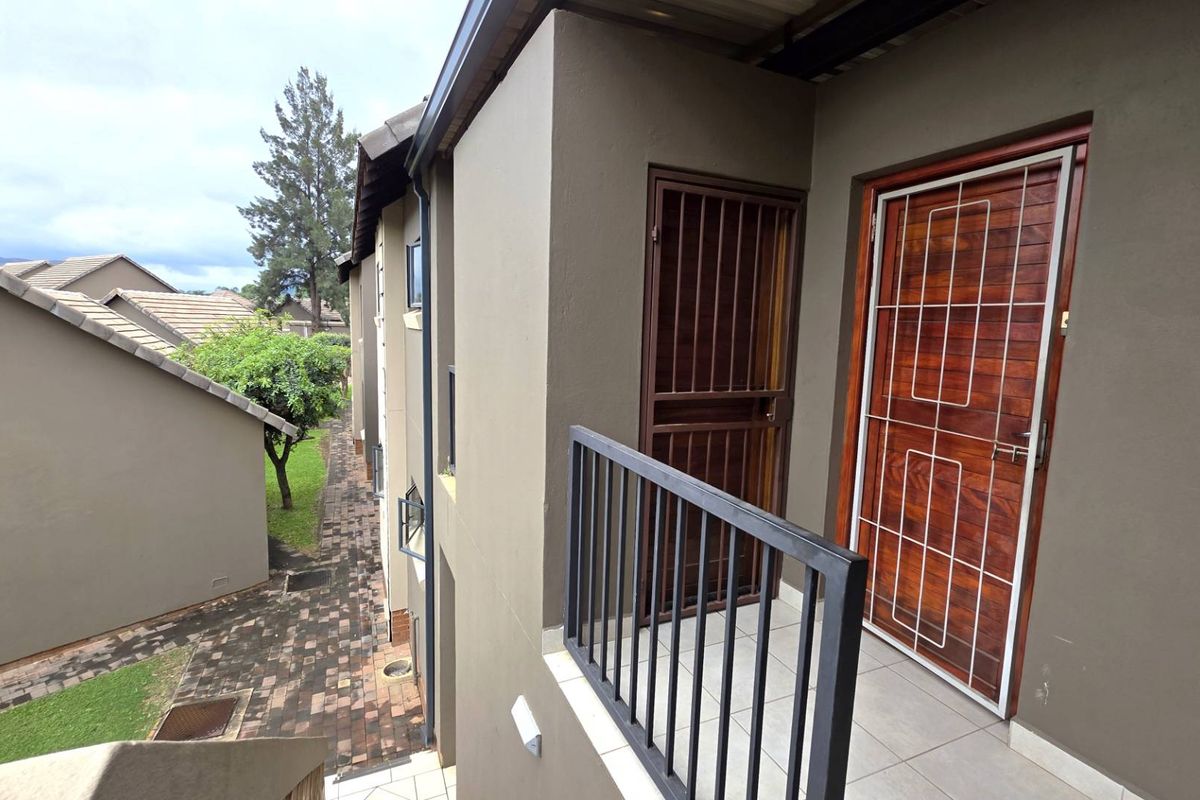 Exterior view of an apartment balcony featuring a security gate, tiled flooring, and metal railing, overlooking a paved walkway and garden area.