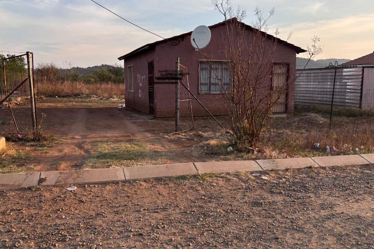 A detached single-story house featuring a basic exterior, security bars, and a satellite dish, situated on an unpaved lot with perimeter fencing.