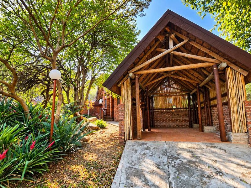 A rustic outdoor entertainment area featuring a thatched roof, timber pole construction, and brick accents, set within a lush, landscaped garden environment.