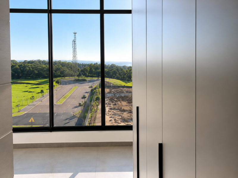 A bright, modern bedroom featuring floor-to-ceiling windows with expansive views, sleek built-in cupboards, and neutral tiled flooring.