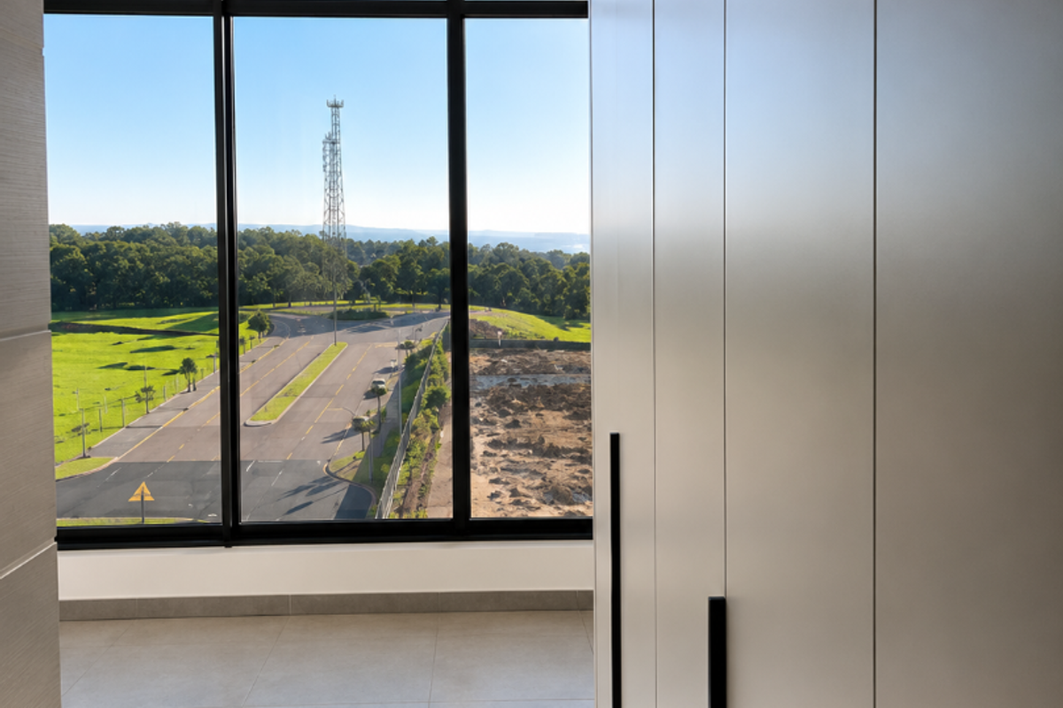A bright, modern bedroom featuring floor-to-ceiling windows with expansive views, sleek built-in cupboards, and neutral tiled flooring.