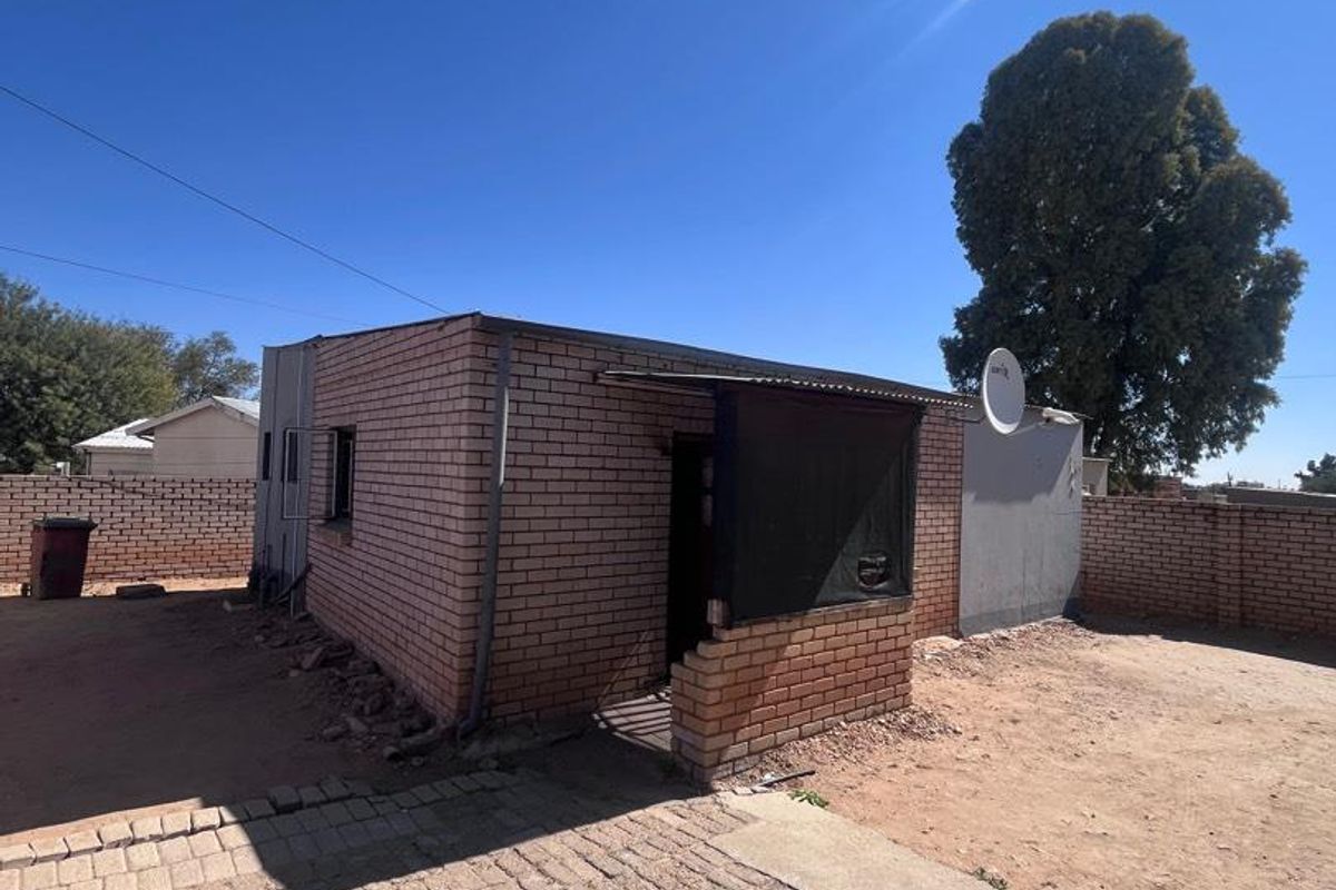 Rear exterior view of a brick house featuring a paved area, boundary walls, and a covered entrance with visible masonry damage.