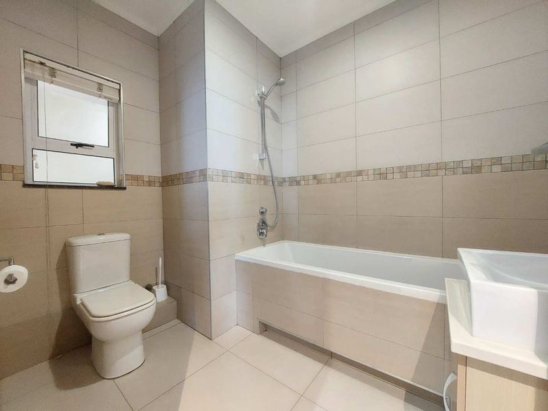 A clean, neutral-toned bathroom featuring a built-in bathtub with shower mixer, a modern toilet, and a vanity basin with tiled finishes.