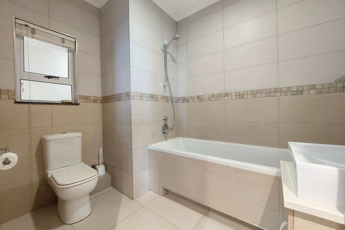 A clean, neutral-toned bathroom featuring a built-in bathtub with shower mixer, a modern toilet, and a vanity basin with tiled finishes.