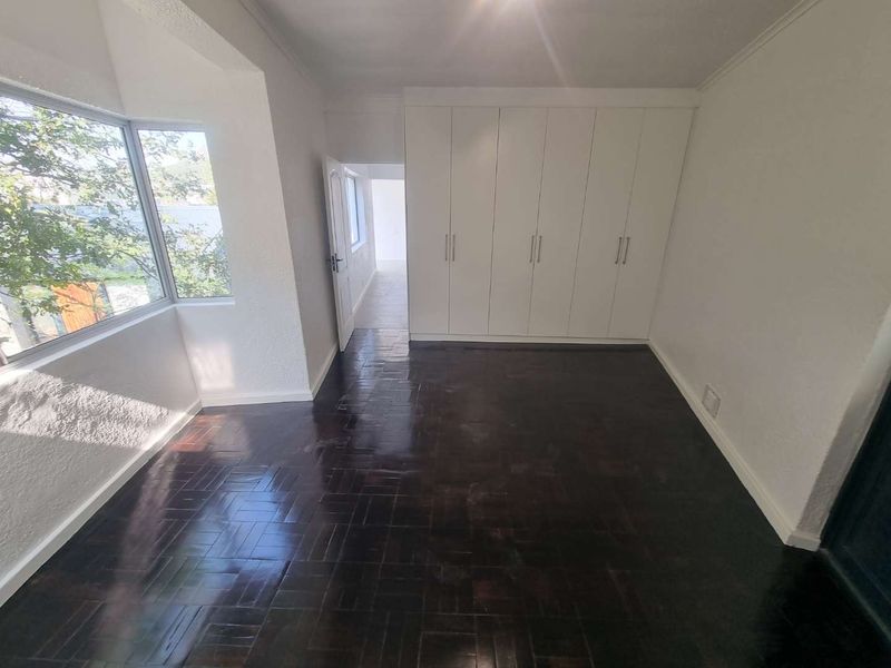 A bright, empty bedroom featuring polished dark parquet flooring, a large bay window, and extensive built-in cupboard storage.