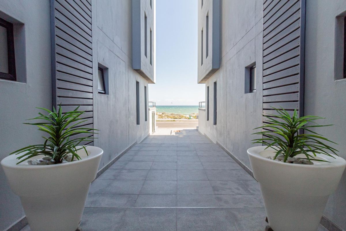 A view through a corridor between apartment buildings, showcasing modern architectural lines and a clear sightline toward the ocean.