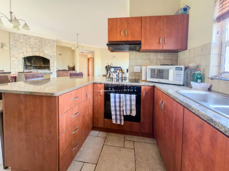 Open-plan kitchen featuring granite countertops, wooden cabinetry, and a built-in oven, flowing into a living area with a stone-clad indoor braai.