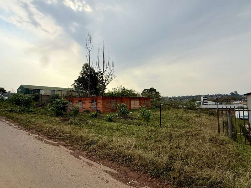 Vacant commercial land featuring a derelict brick structure with boarded windows, enclosed by metal palisade fencing and overgrown vegetation.