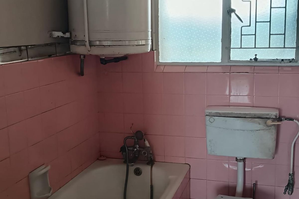 A dated bathroom featuring pink wall tiles, a bathtub, a toilet with a wall-mounted cistern, and an exposed electric water heater.