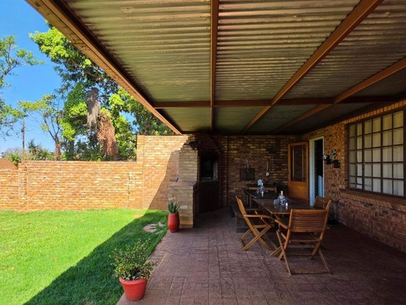 Covered outdoor patio featuring a built-in brick braai, corrugated metal roofing, and paved flooring, overlooking a private garden area.