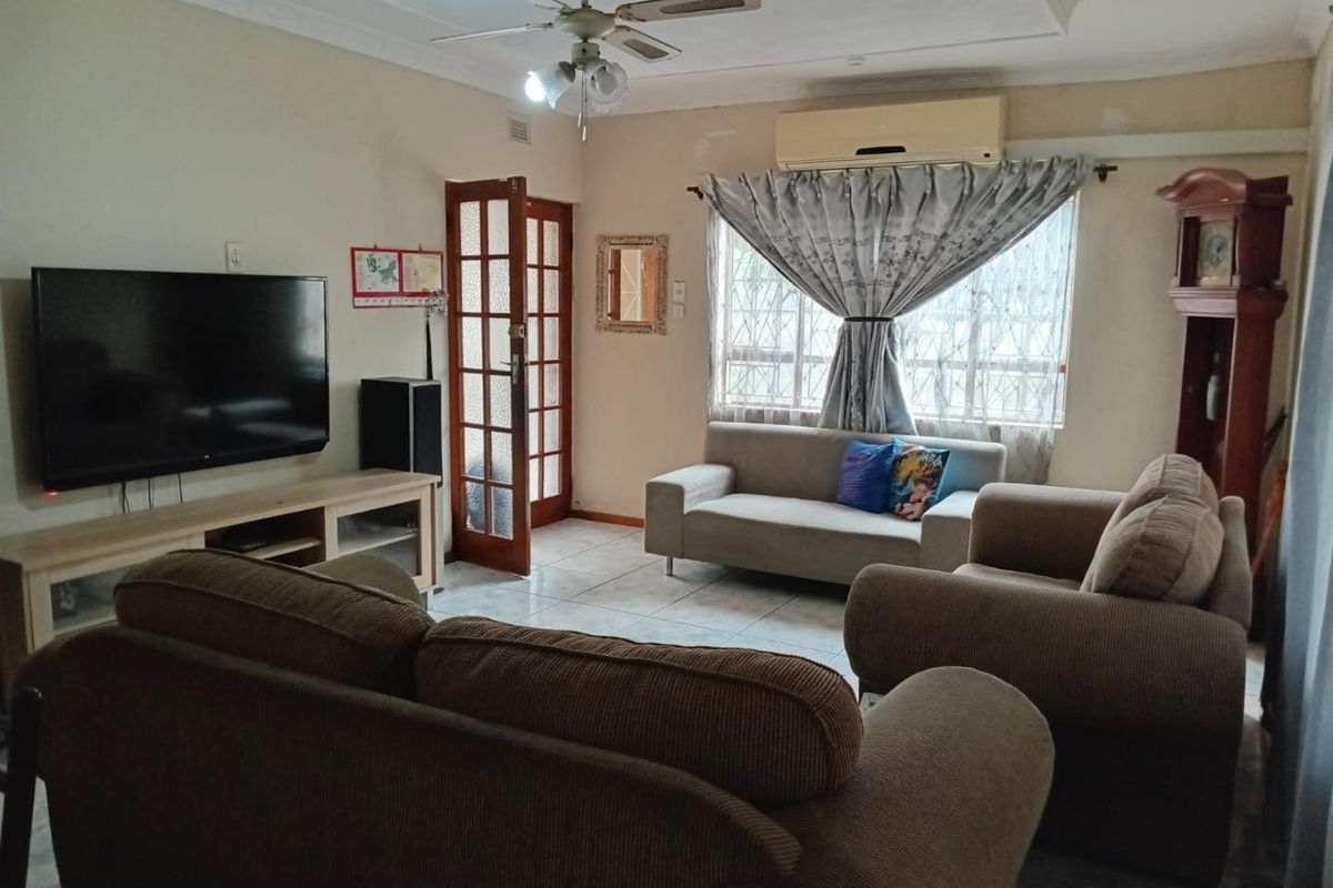 A comfortable living room featuring tiled flooring, a ceiling fan, and a wall-mounted split-system air conditioner, with natural light through a security-barred window.