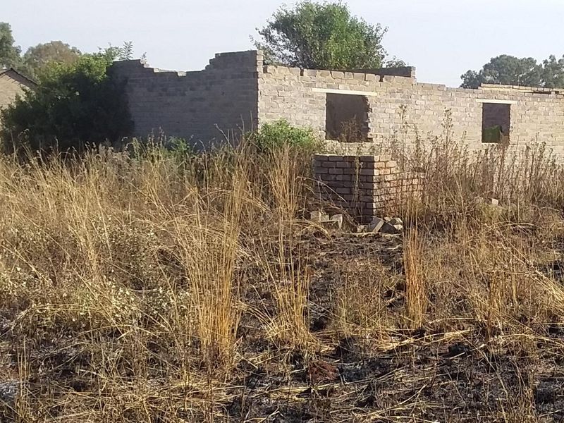 A view of a vacant residential plot featuring an incomplete brick structure with no roof, set within an overgrown, undeveloped landscape.