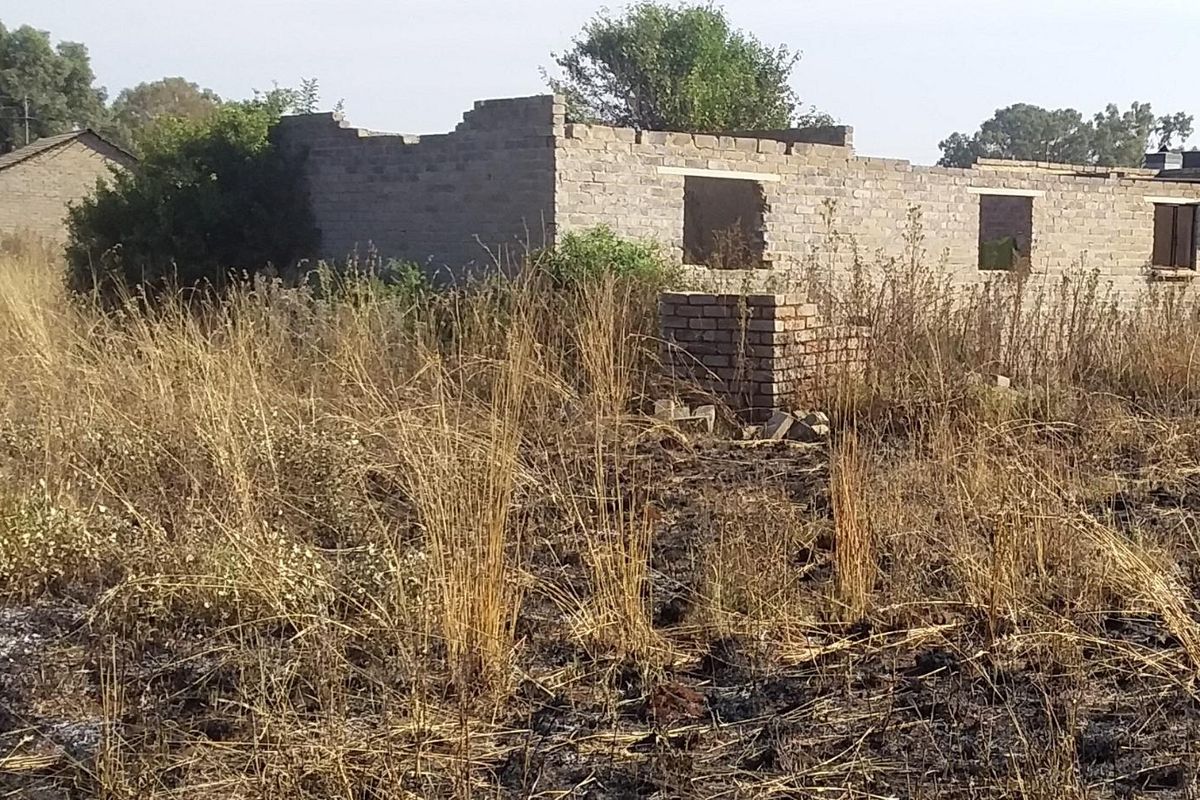 A view of a vacant residential plot featuring an incomplete brick structure with no roof, set within an overgrown, undeveloped landscape.