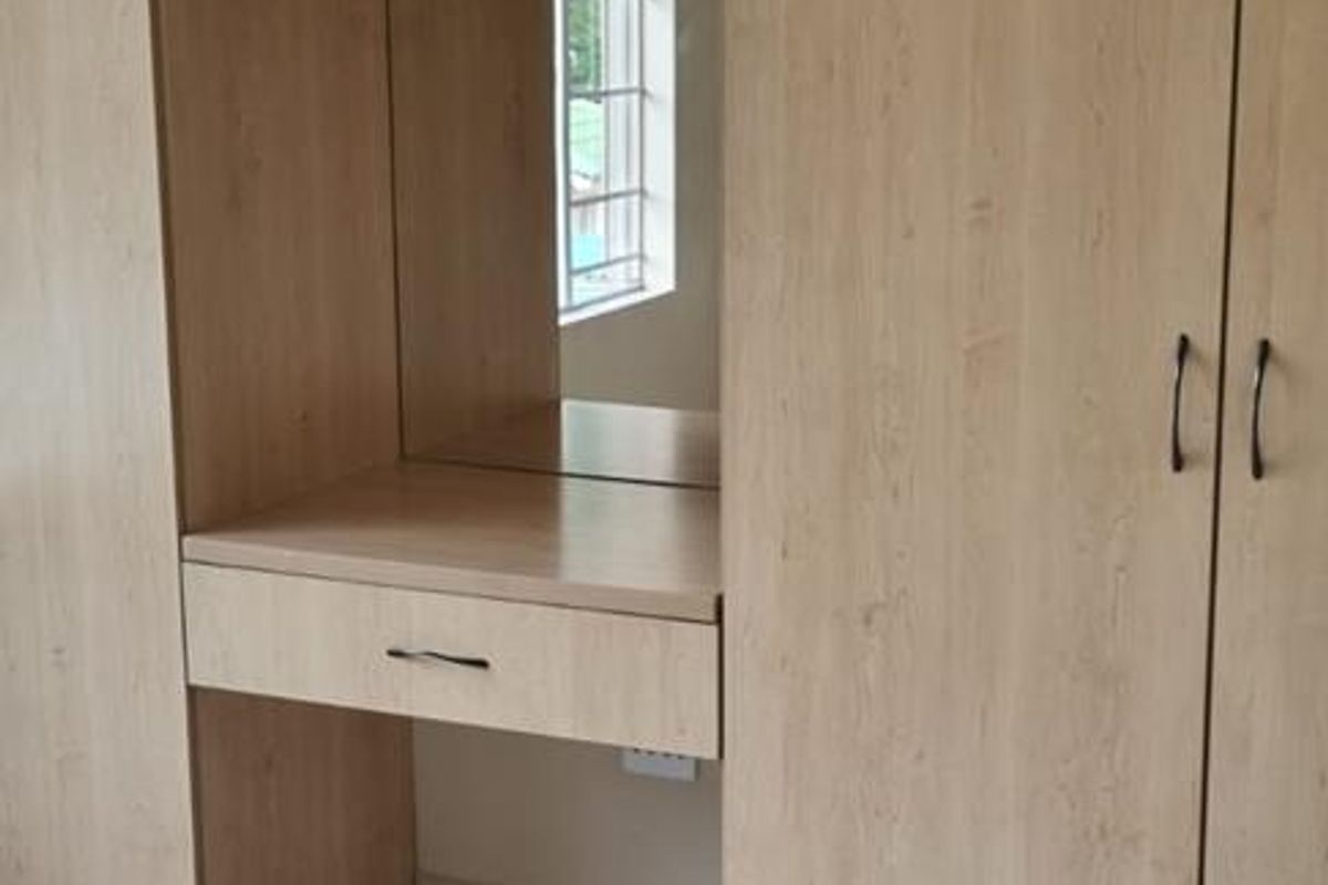 A bedroom featuring light wood-toned built-in wardrobes with an integrated vanity station and mirror, complemented by neutral tiled flooring.