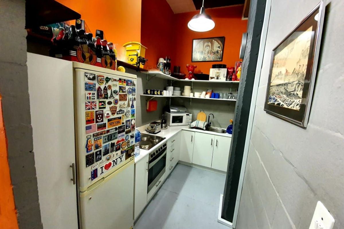 A compact office kitchen featuring white cabinetry, a built-in oven, cooktop, stainless steel sink, and open shelving against a vibrant orange wall.