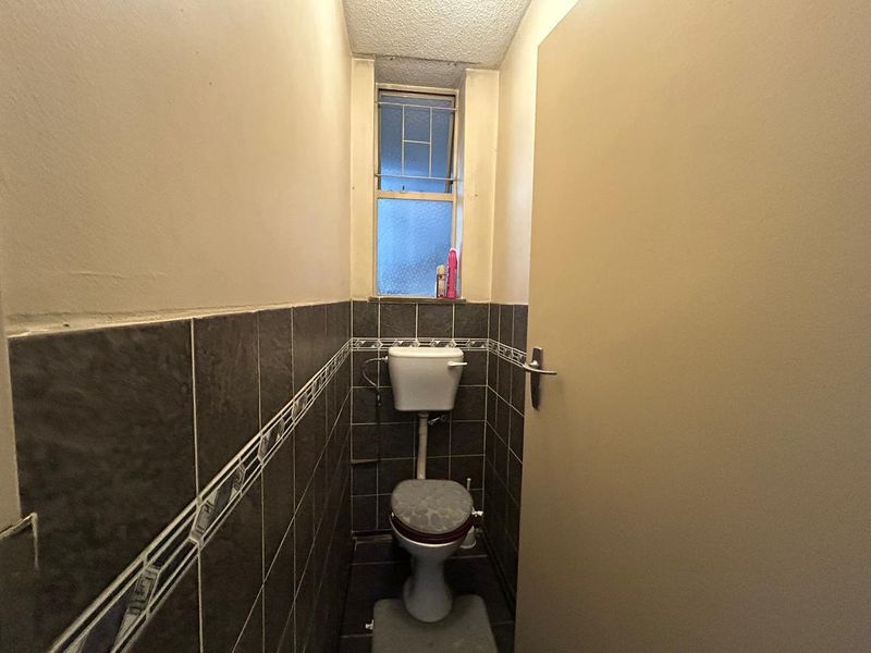 A compact, dated bathroom featuring dark wall tiles, a standard toilet, and a small window providing natural light.