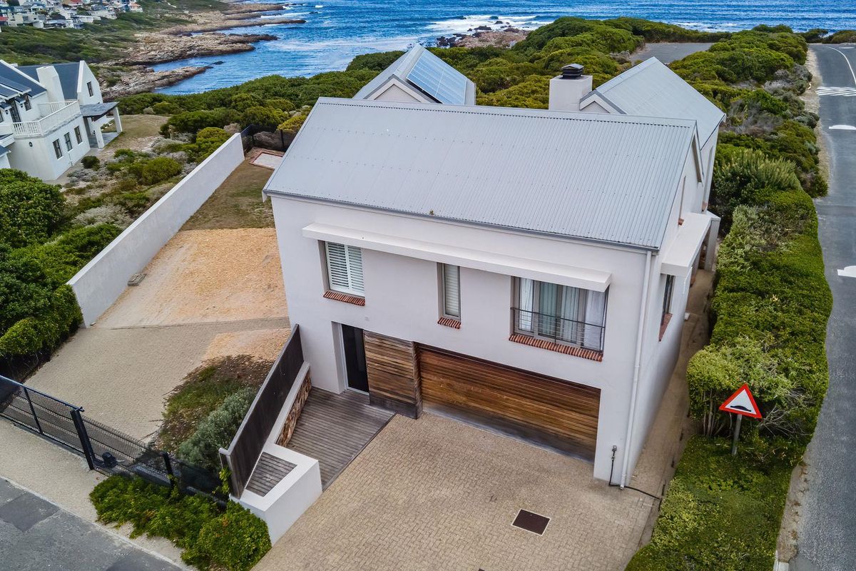 Aerial view of a modern coastal residence featuring a metal roof, solar panels, timber garage door, and paved driveway near the ocean.