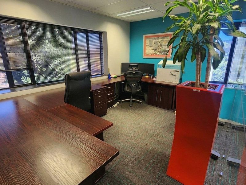 A well-lit office space featuring large windows with venetian blinds, carpeted flooring, and a modern glass partition wall.